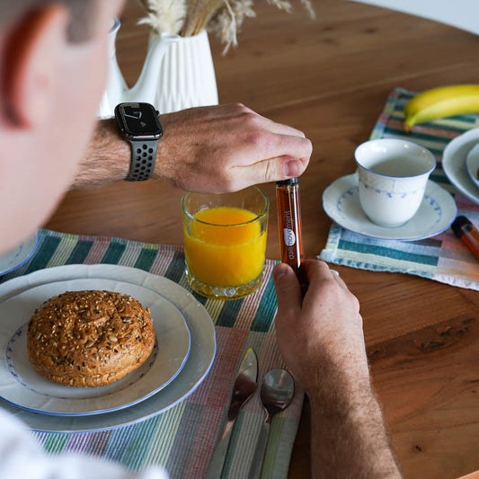 Persoon die aan de ontbijttafel een vloeibaar supplement inneemt naast een glas sinaasappelsap en een broodje.