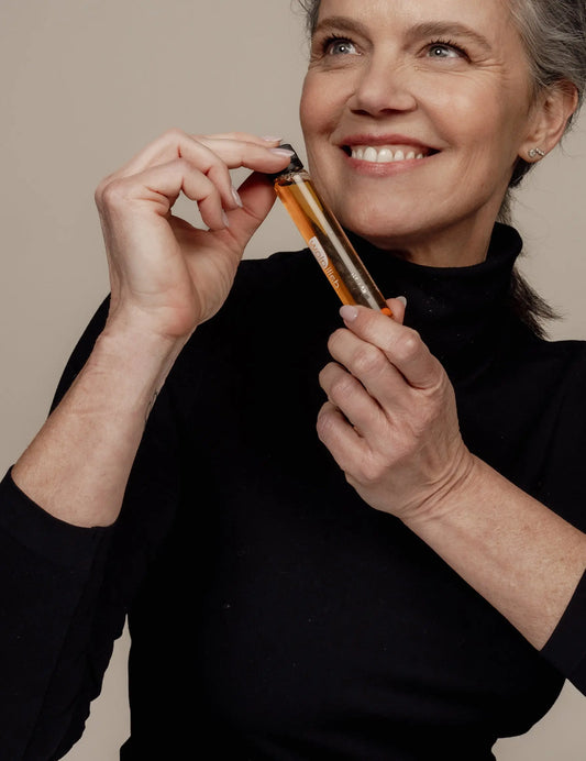 Vrouw met grijs haar bekijkt een kleine glazen ampul in haar hand in een rustige studio-omgeving.