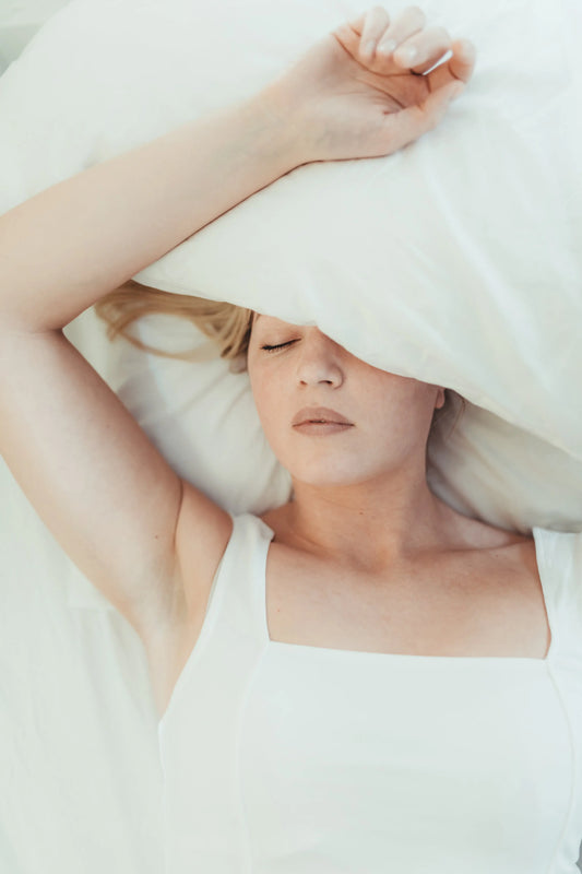 Vrouw ligt ontspannen in bed met gesloten ogen en een kussen deels over haar hoofd, symbool voor rust en slaap.
